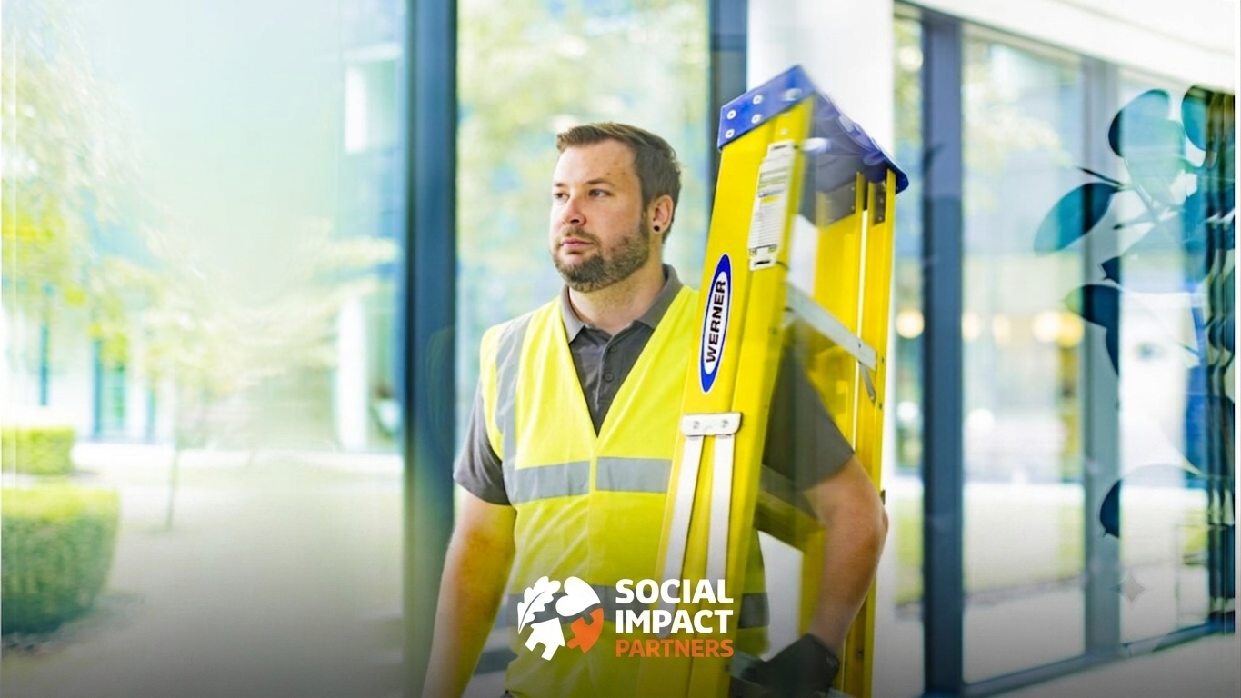 A person wearing a high-visibility yellow safety vest stands beside a yellow extension ladder in a modern building with large glass windows. The bright indoor setting features natural light and reflections on the glass. The Social Impact Partners logo appears at the bottom of the image.