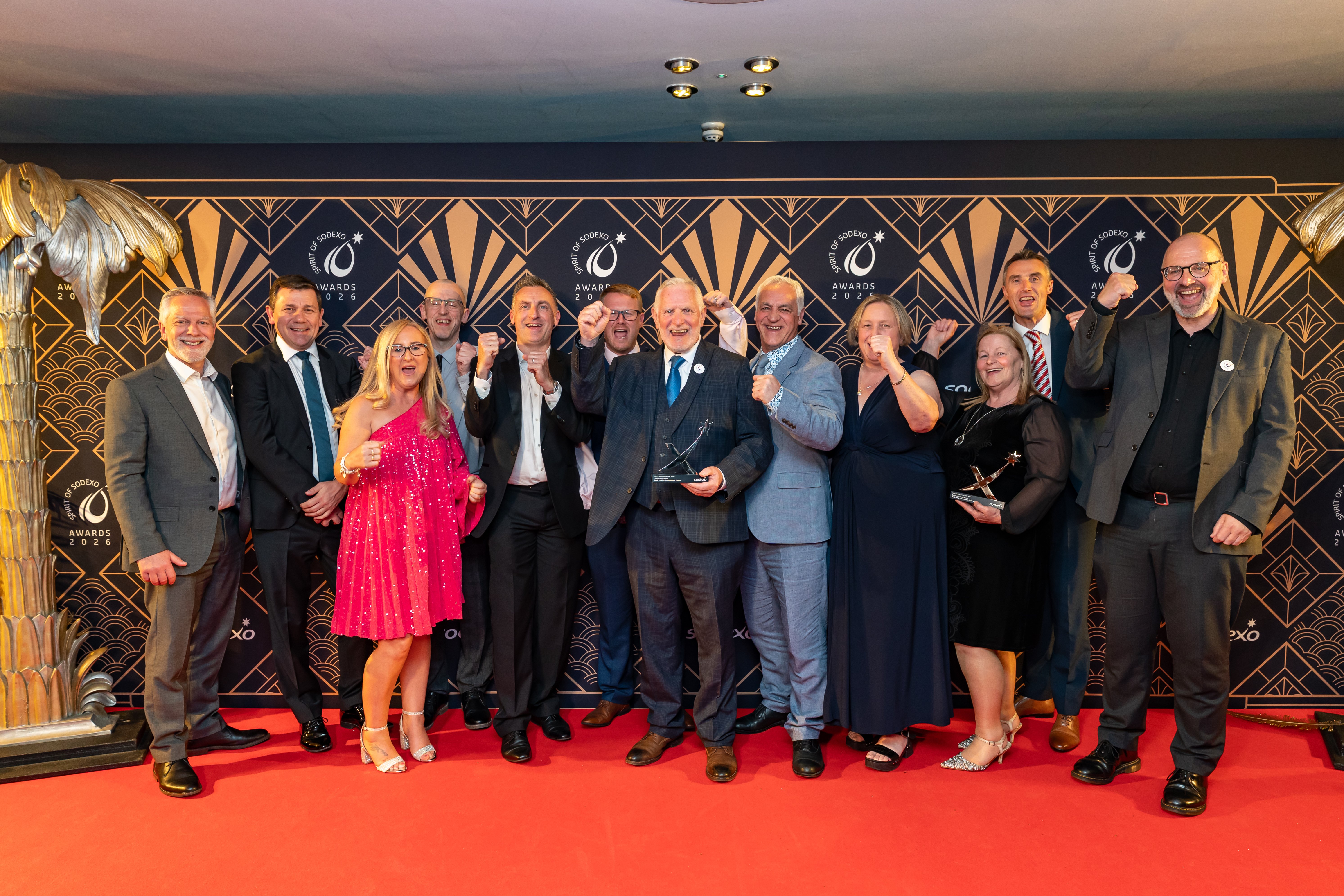 Group of formally dressed attendees posing on a red carpet at the Spirit of Sodexo Awards 2026 backdrop, several holding glass trophies and raising their fists, with an art deco–style black and gold branded wall behind them.