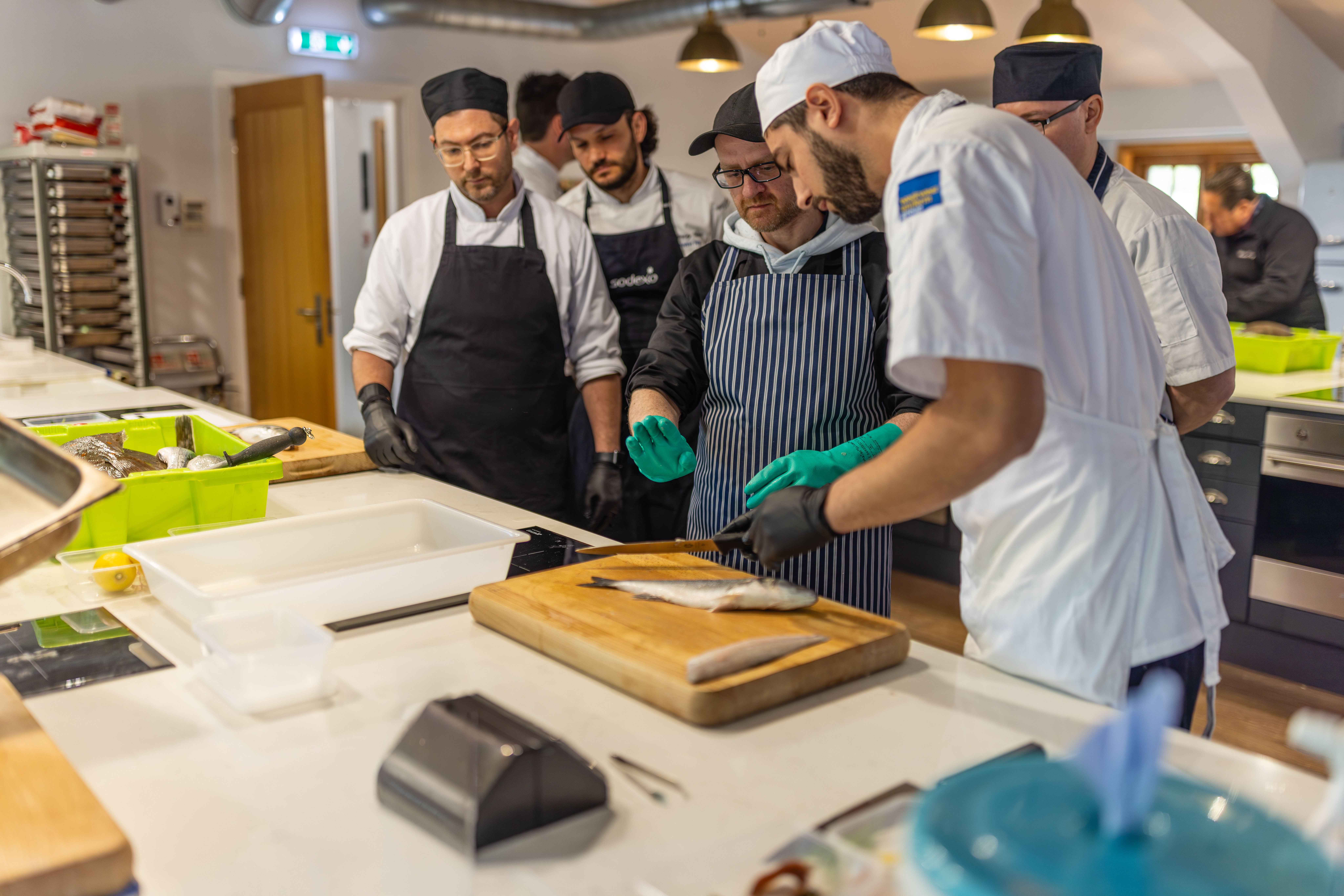 Commercial kitchen setting where several people in chef uniforms and aprons are gathered around a work surface. In the foreground, one person wearing blue gloves is preparing food at a cutting board with utensils and containers nearby, while others stand behind observing and engaging in the activity.