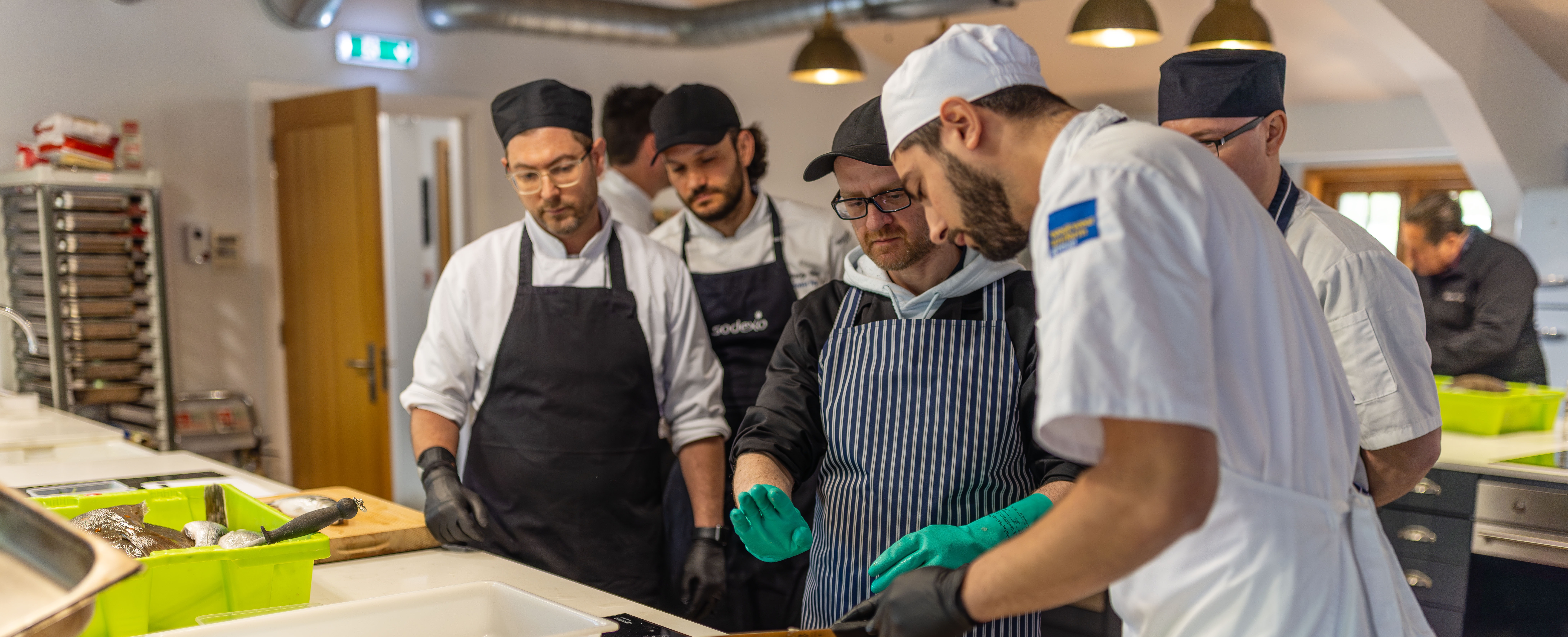 Commercial kitchen setting where several people in chef uniforms and aprons are gathered around a work surface. In the foreground, one person wearing blue gloves is preparing food at a cutting board with utensils and containers nearby, while others stand behind observing and engaging in the activity.