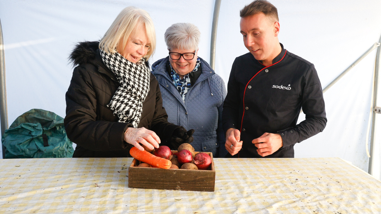Two women and a man in a chef uniform examining a crate of fresh vegetables.