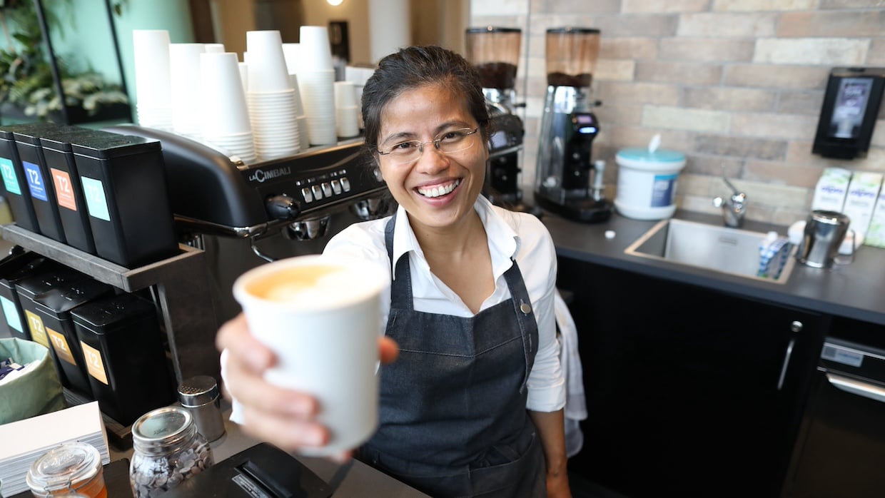 A female café worker is smiling and holding up a cup filled with coffee. 