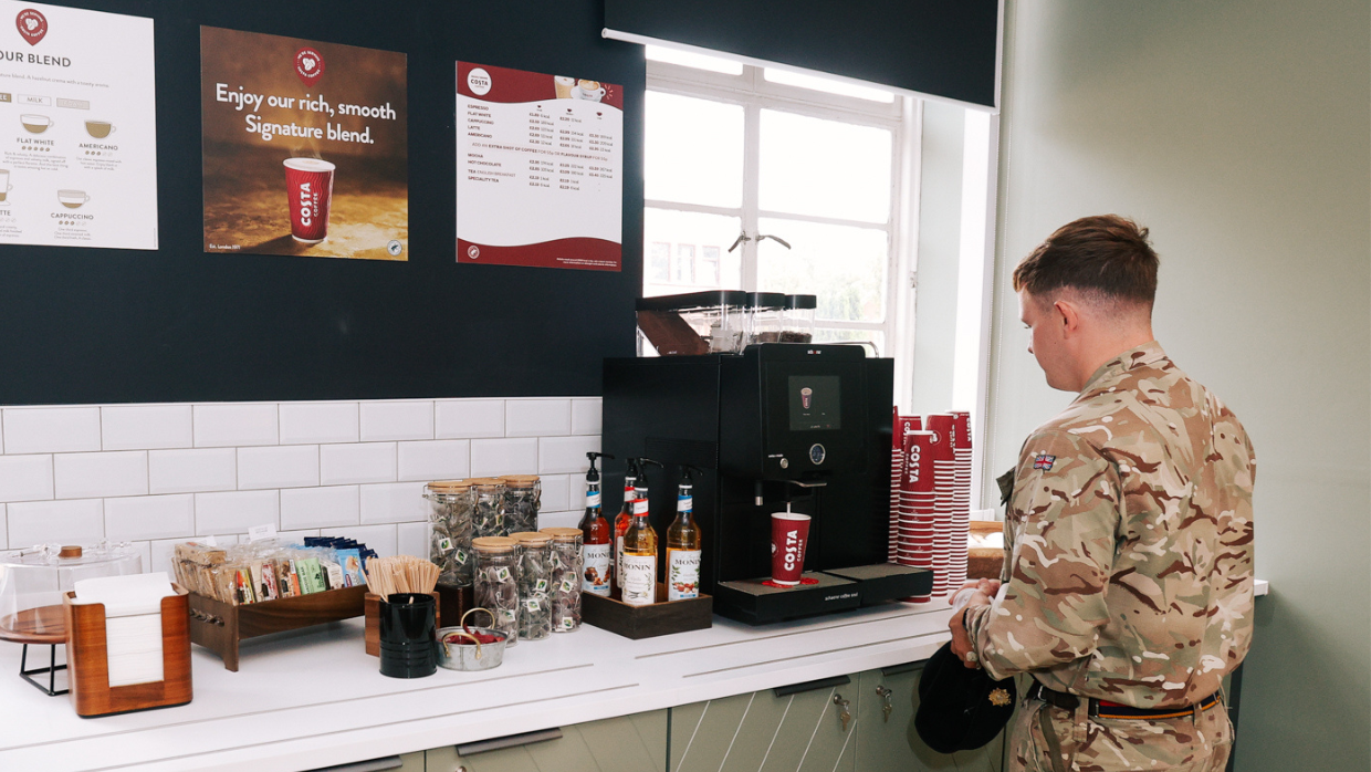 A soldier in uniform stands at a counter, getting a hot drink at a coffee machine.