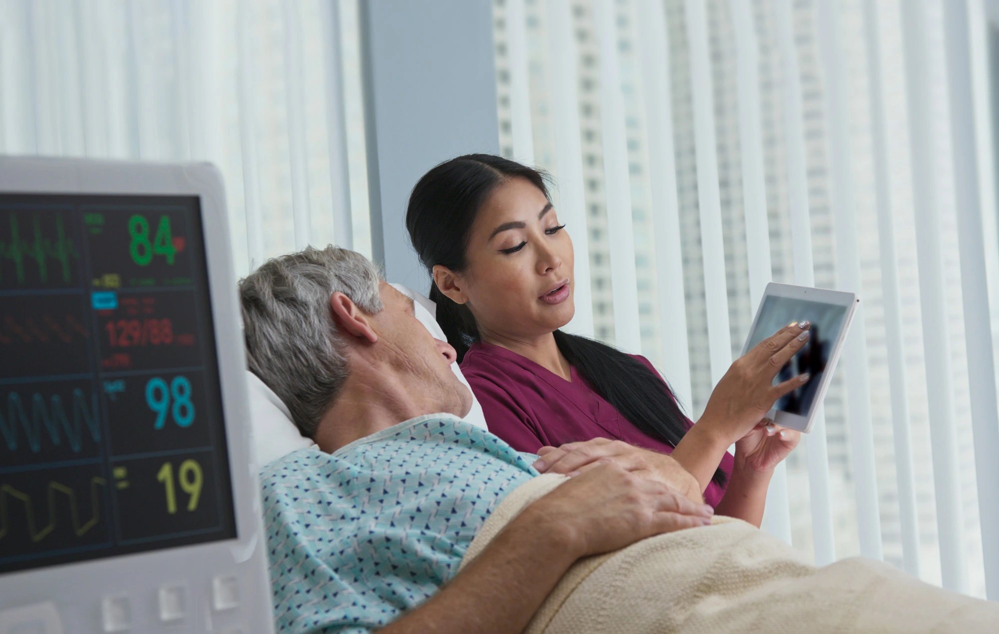 Nurse showing a hospital inpatient how to use a tablet, with vital signs monitor displayed beside the bed.