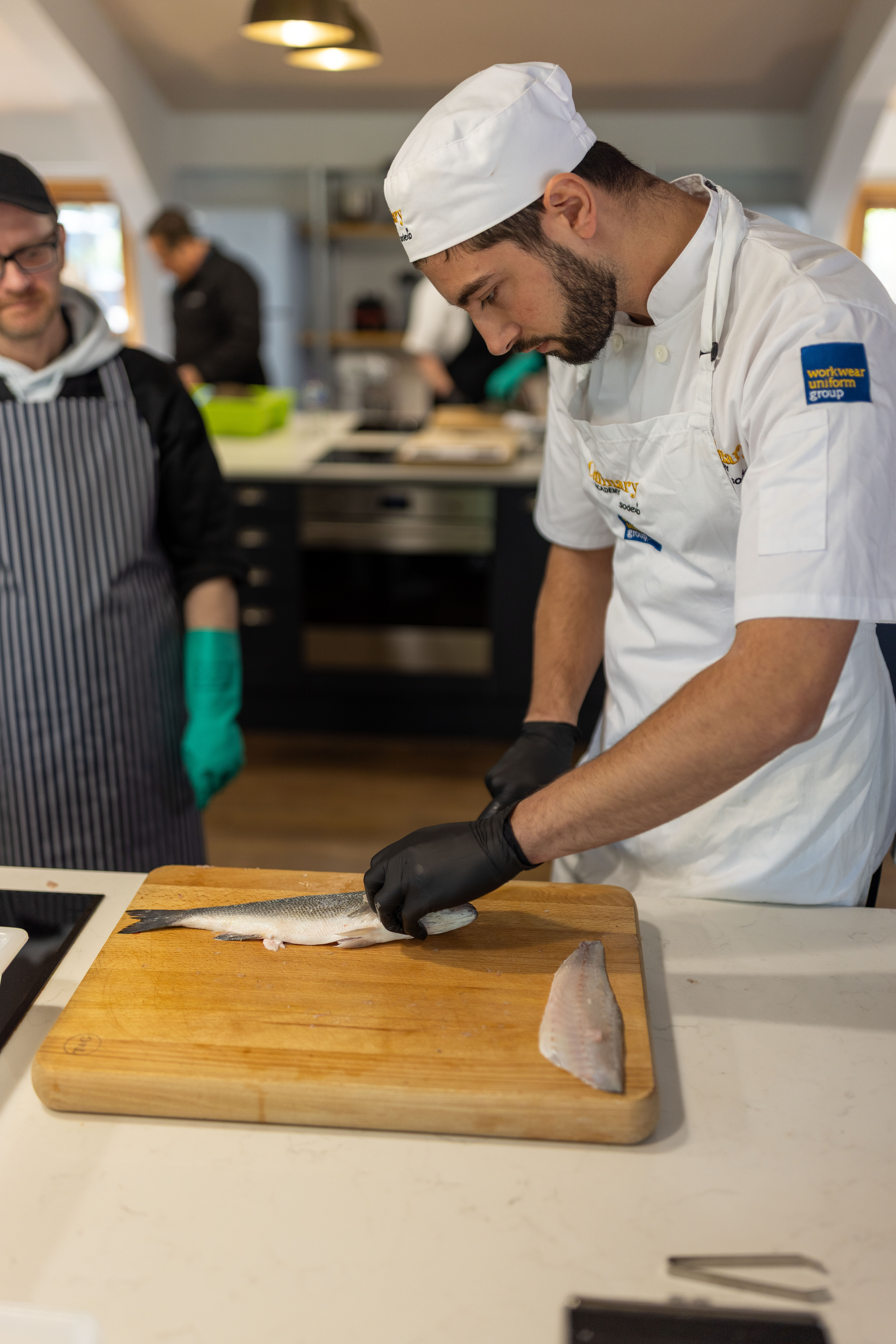 Commercial kitchen scene showing a person in a white chef uniform and grey cap working at a wooden cutting board. The person is using a handheld kitchen tool to prepare ingredients, with blue gloves placed nearby. Stainless steel appliances and other people in chef attire are visible in the softly blurred background.