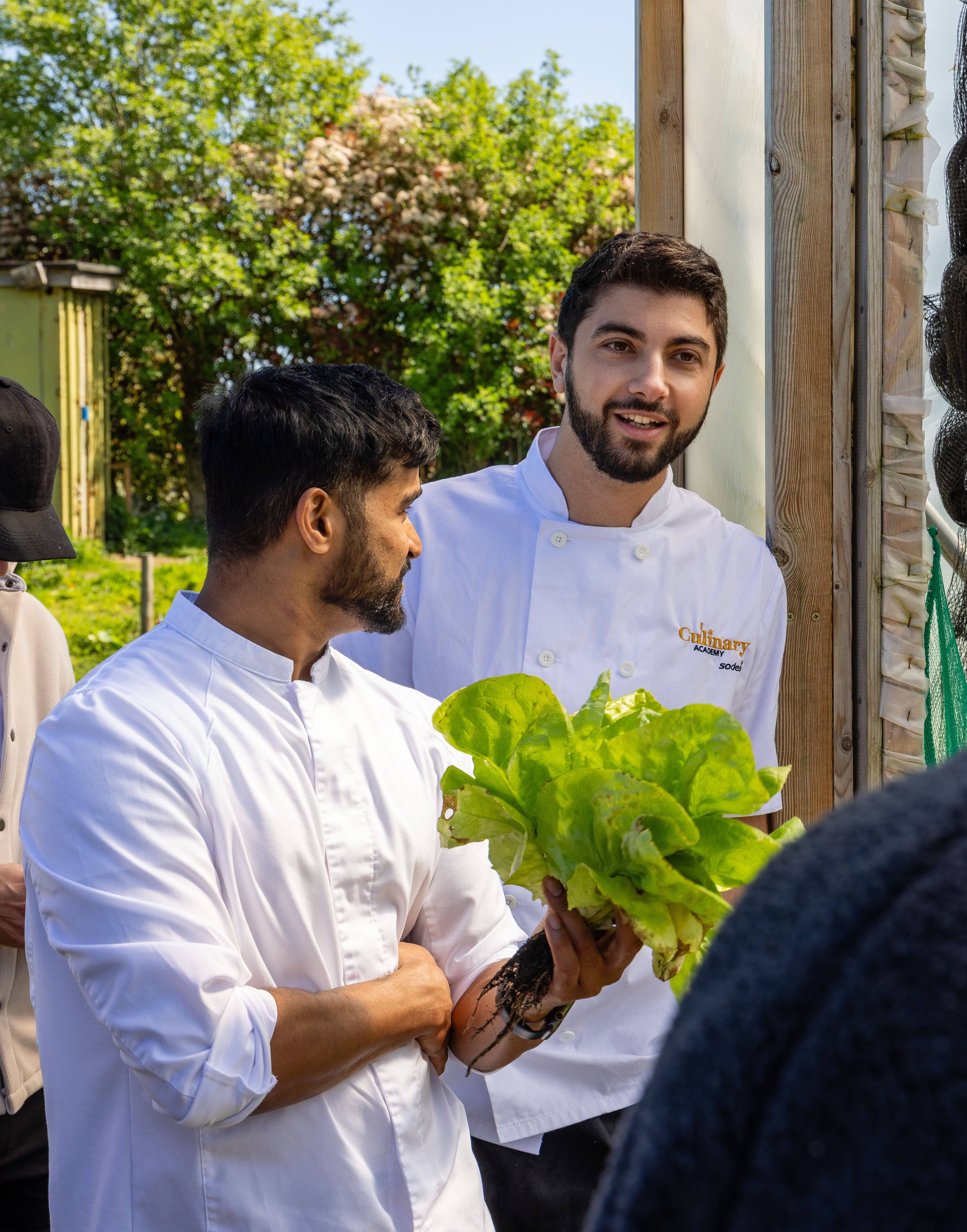 Two people standing by an open doorway in an outdoor setting. One person holds a bunch of fresh leafy greens while speaking to another person wearing a white chef-style jacket. Greenery and plants are visible in the background, suggesting a garden or farm environment.