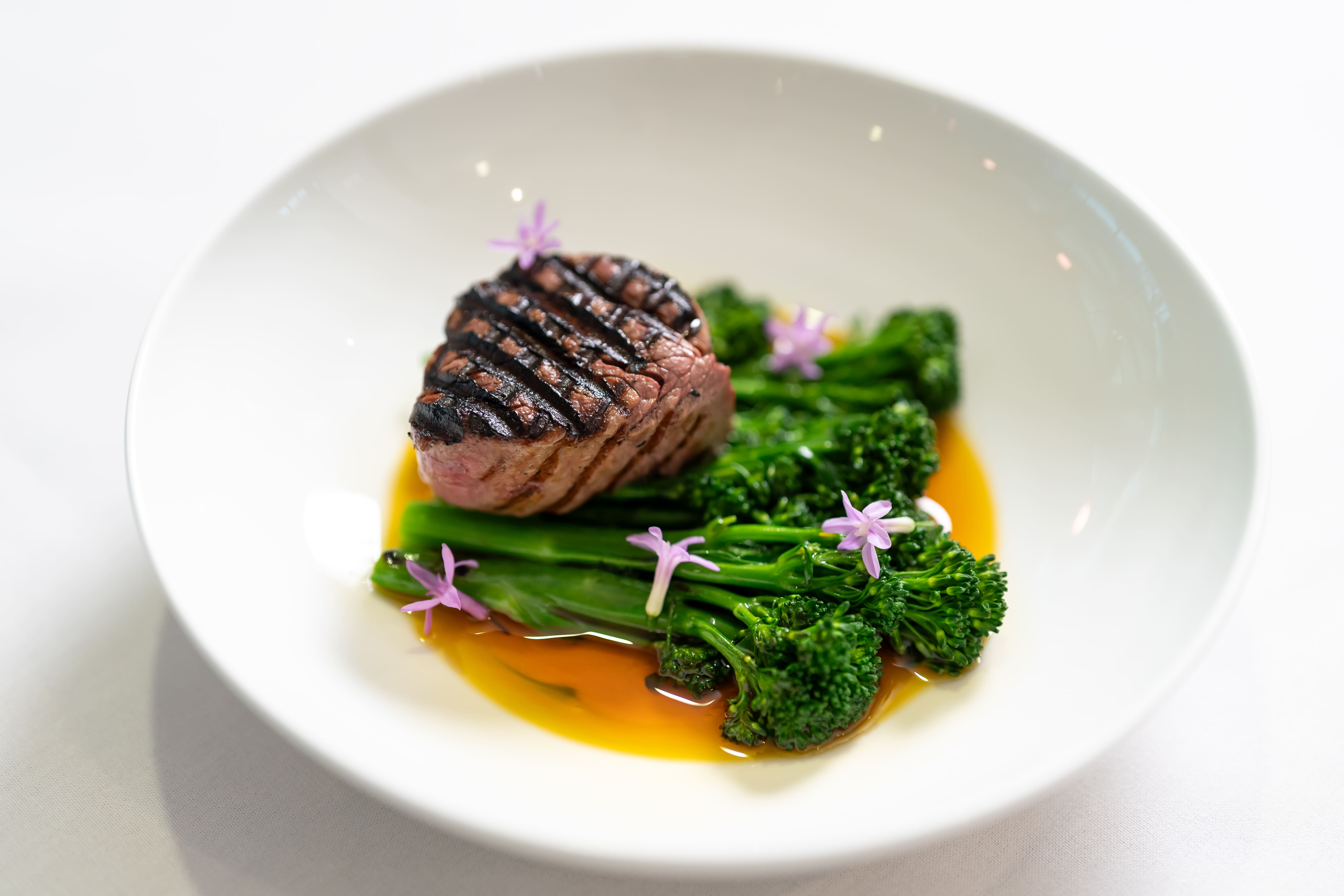 A plated dish featuring a grilled piece of meat served on broccolini with edible flowers and sauce.