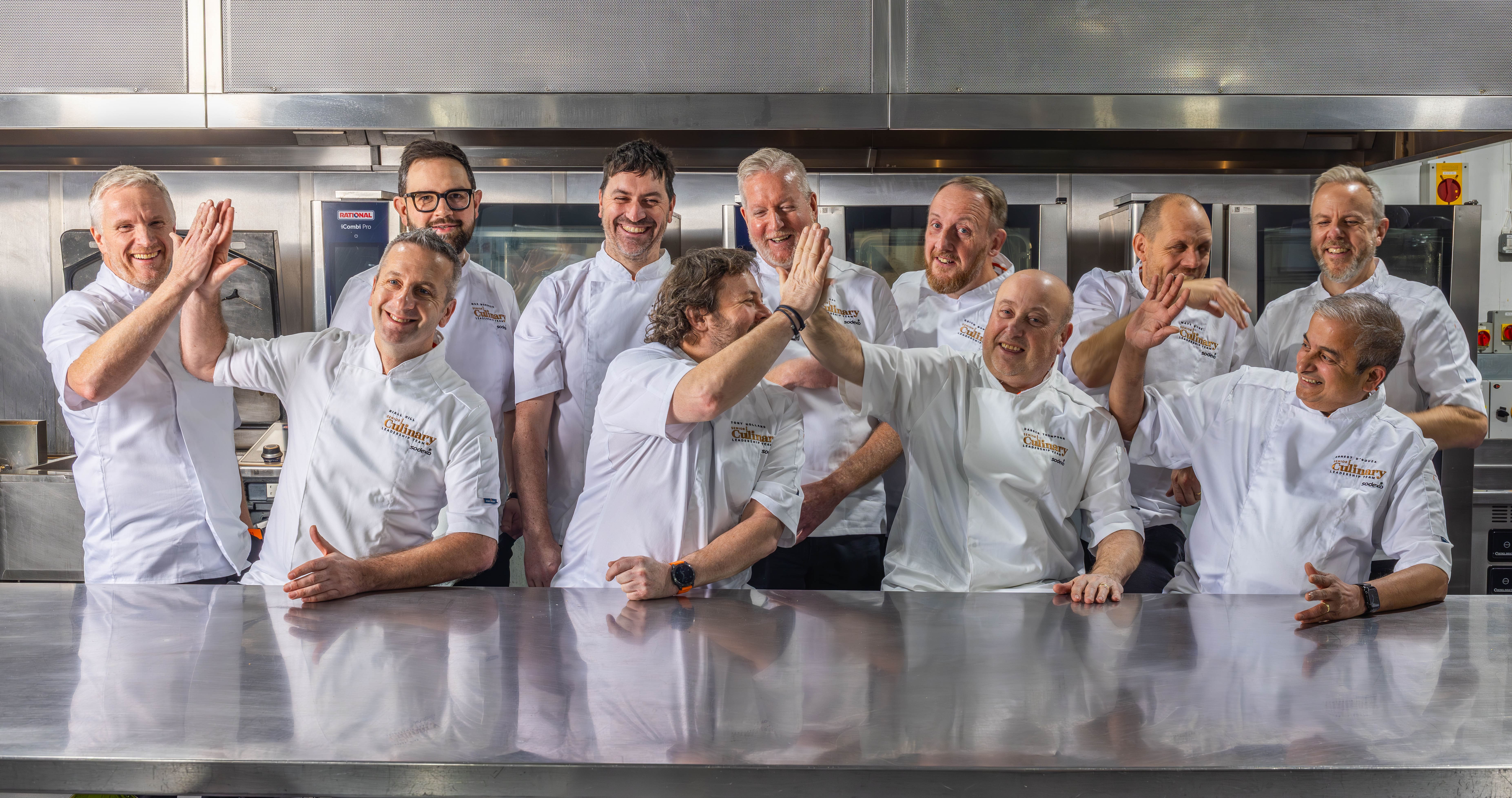A group of chefs in white uniforms standing together in a commercial kitchen, raising their hands in a lively moment.