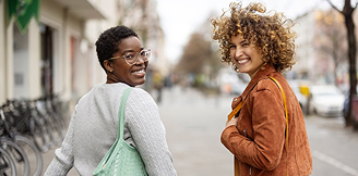 Two people walking along a city street, talking while carrying bags.