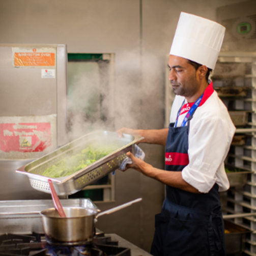 A chef with white hat pouring hot veg into a pan