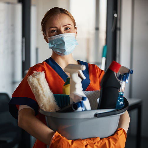 A woman wearing a face mask holding a bucket of cleaning supplies