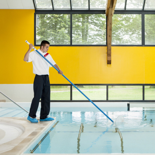 A man in swim trunks using a pool net to clean leaves and debris from the surface of a swimming pool