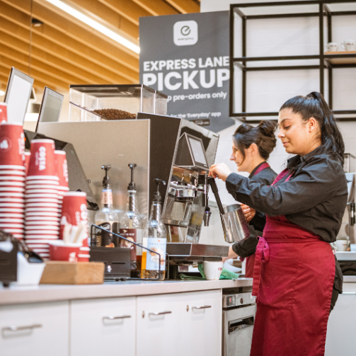 Woman in apron standing behind counter taking a coffee