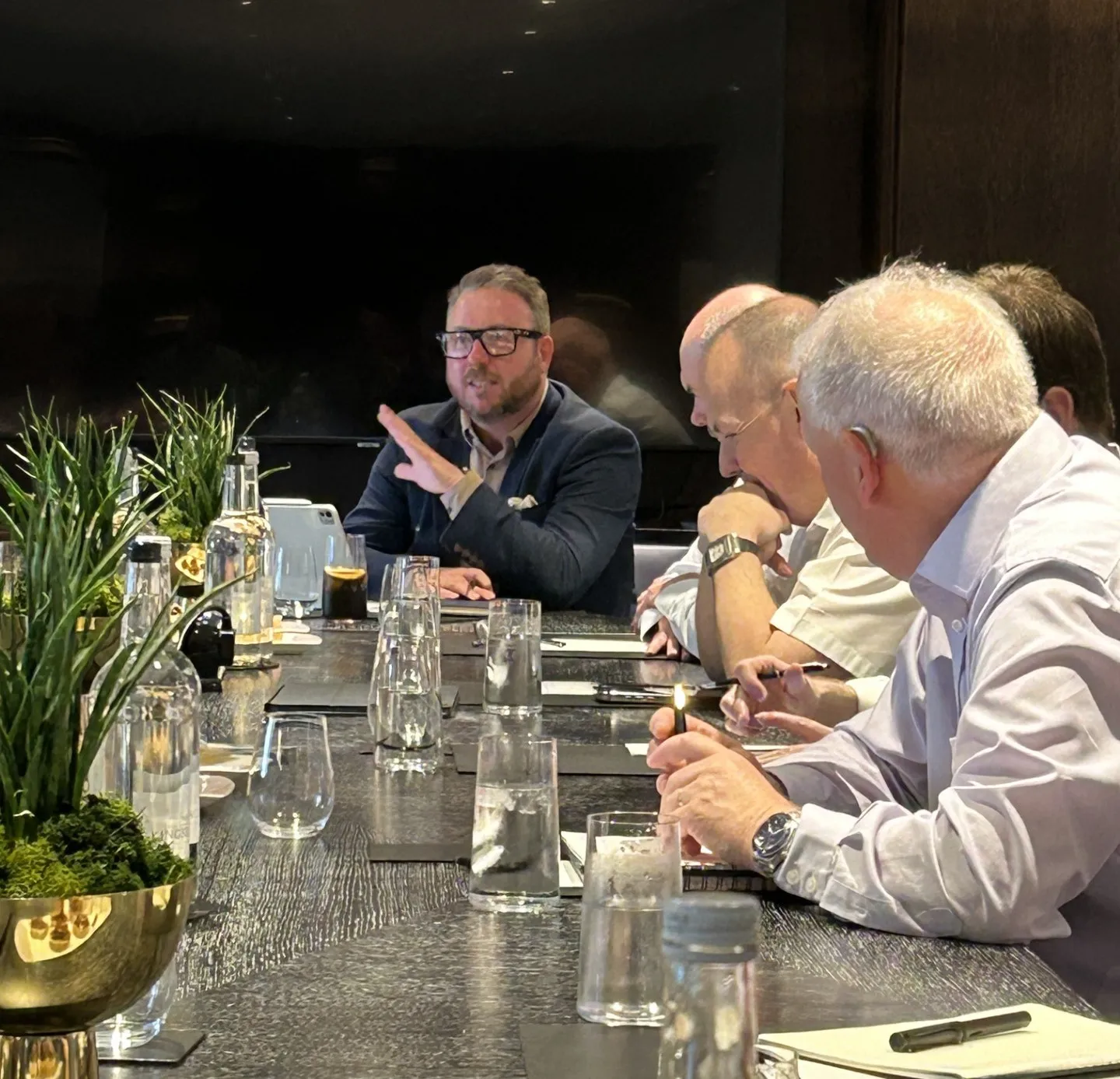 Participants seated around a conference table during a roundtable discussion. One person speaks while gesturing, as others listen and take notes. Water glasses, notebooks and decorative plants are arranged along the table in a modern meeting room.