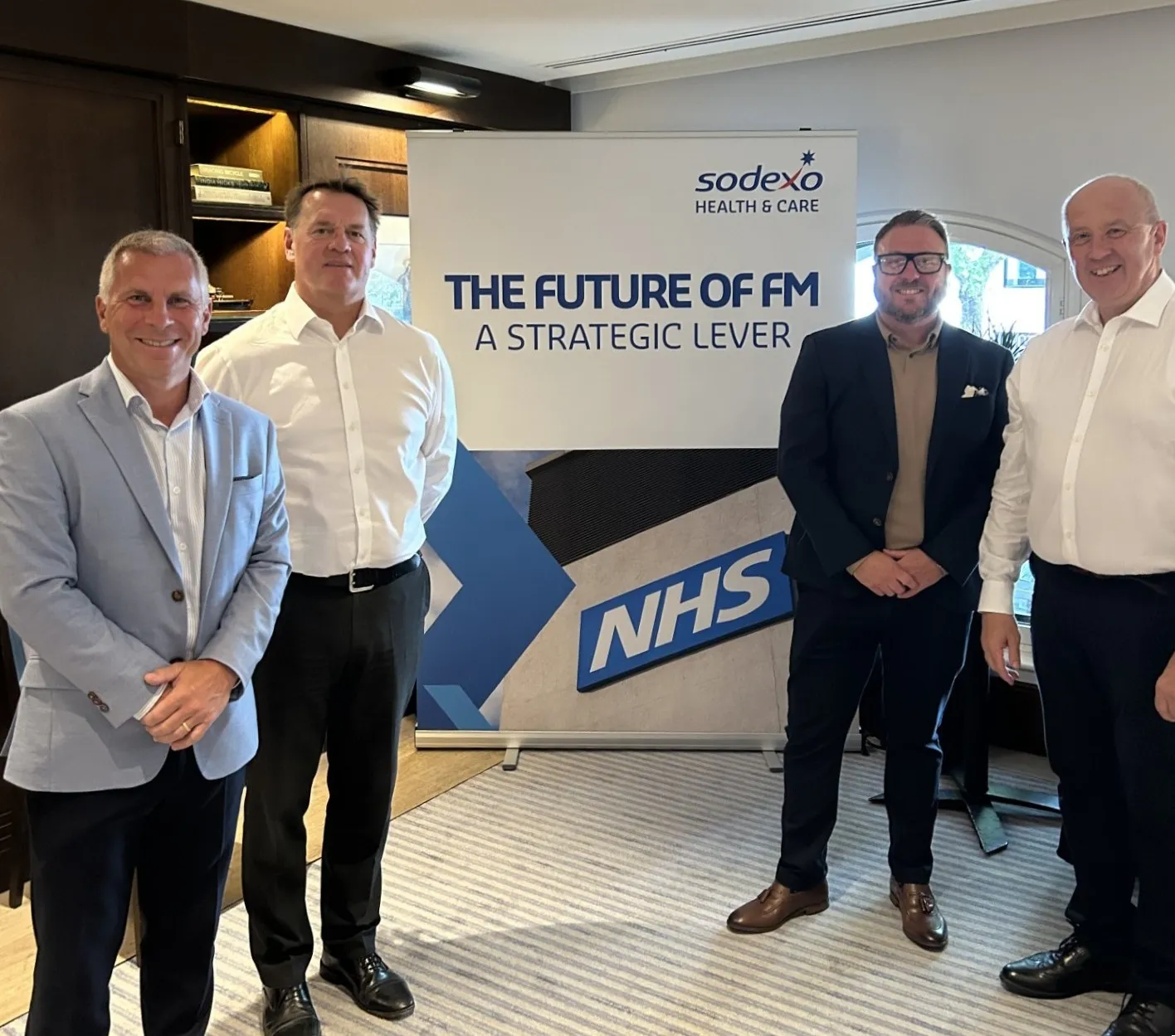 Four attendees stand beside a Sodexo Health & Care banner displaying the text “The Future of FM: A Strategic Lever” and the NHS logo. The group is positioned in a meeting venue with wood-panelled walls and natural light from a nearby window.
