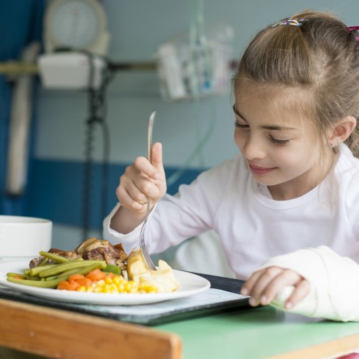A little girl with her arm in a cast is in a hospital bed, holding a fork and eating nutritious food