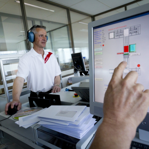 A hand interacting with a screen, there’s a male worker wearing headphones in the background