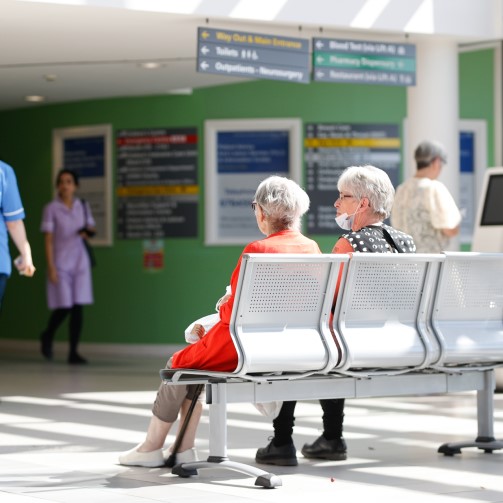Two older patients sat in hospital reception waiting for their appointment as nurses pass by