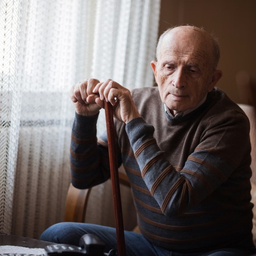 Older male sat in chair, leaning on his walking stick as he attempts to get up out of the chair