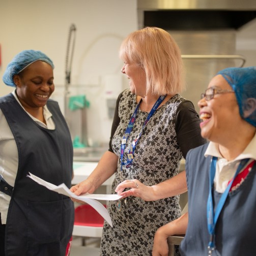 Two hospital kitchen workers are getting instructions from their manager, laughing together