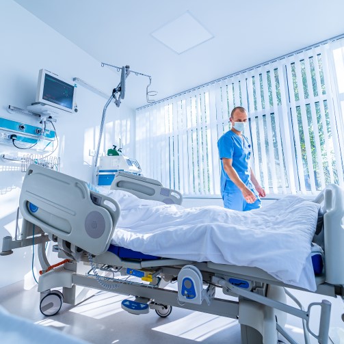 A nurse in uniform wearing a mask is standing next to a hospital bed in a hospital room