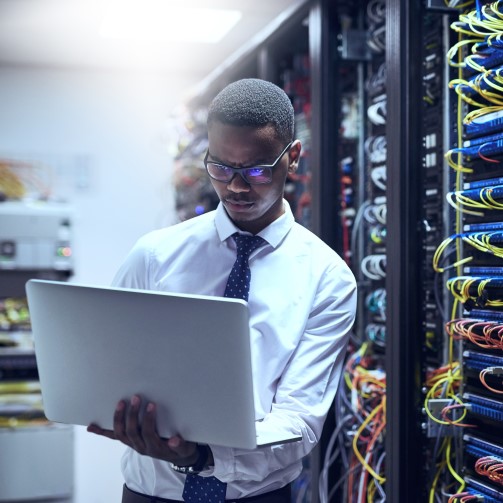 A man is holding a laptop, standing in front of IT cables