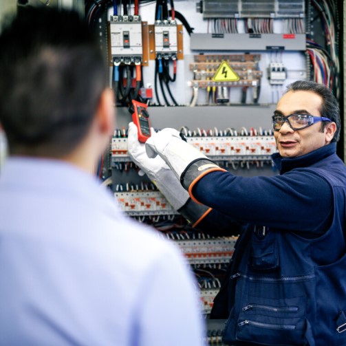 A worker is wearing a pair of goggles and looking at his colleague whilst working on an electric panel