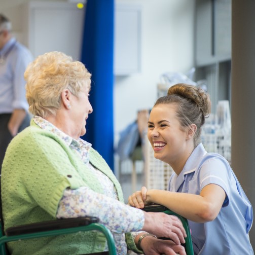 A nurse is kneeled down by a female elderly patient in a wheelchair, both smiling