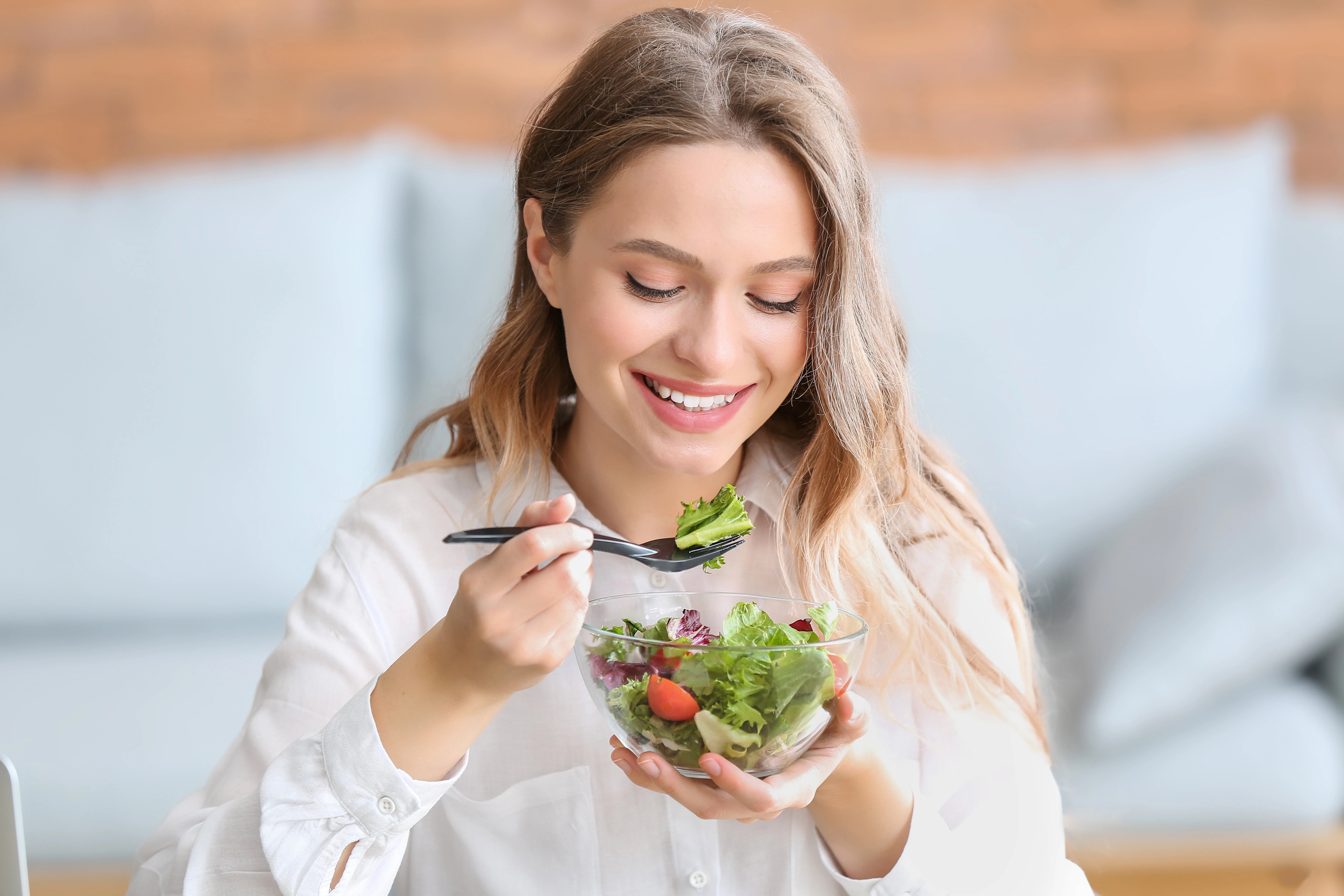 Person holding a bowl of mixed salad and using a fork to eat leafy greens, tomatoes, and vegetables in a bright indoor setting.