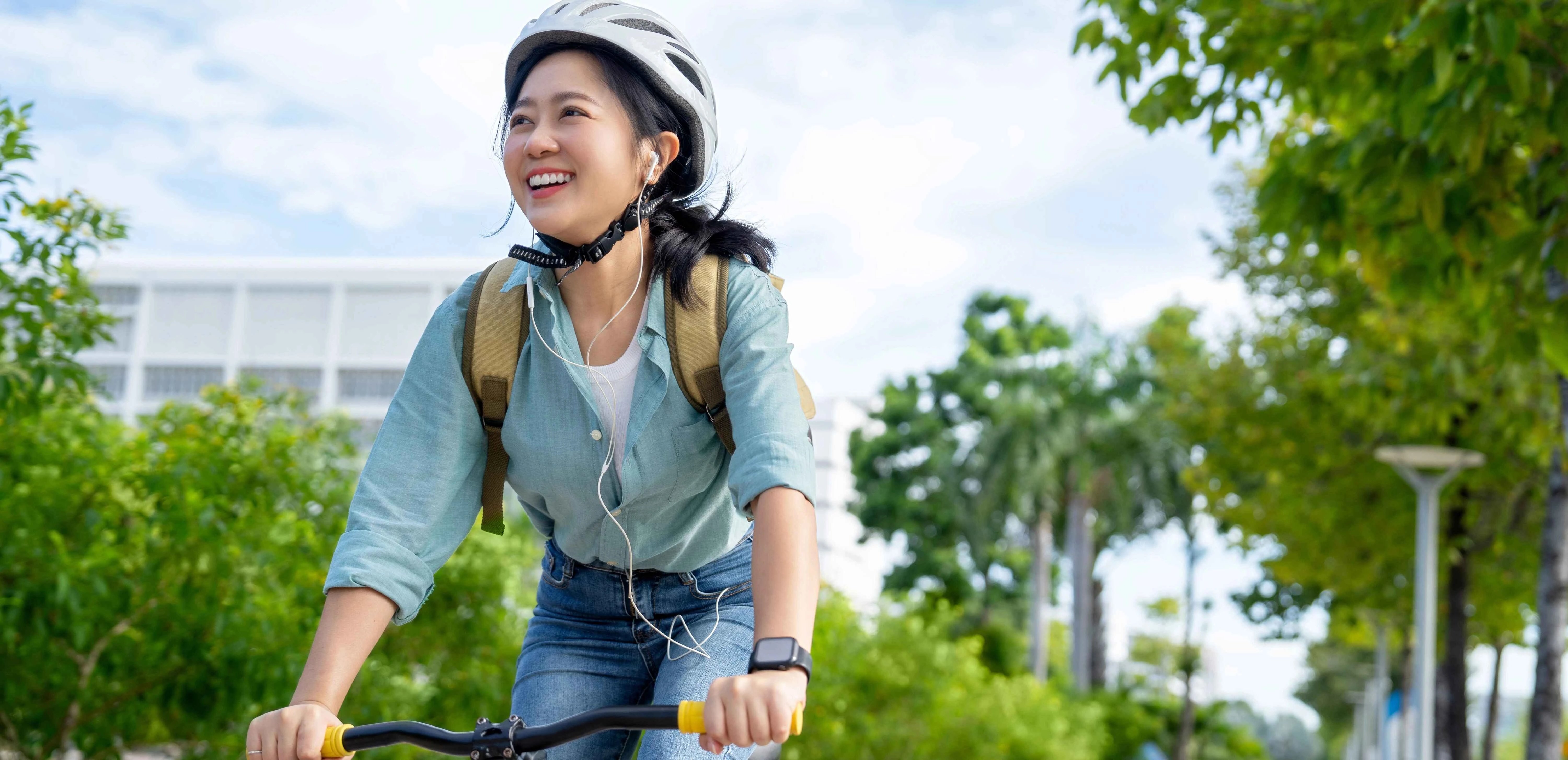 Person riding a bicycle on a tree-lined path while wearing a helmet, backpack, and earphones, with greenery and buildings in the background.
