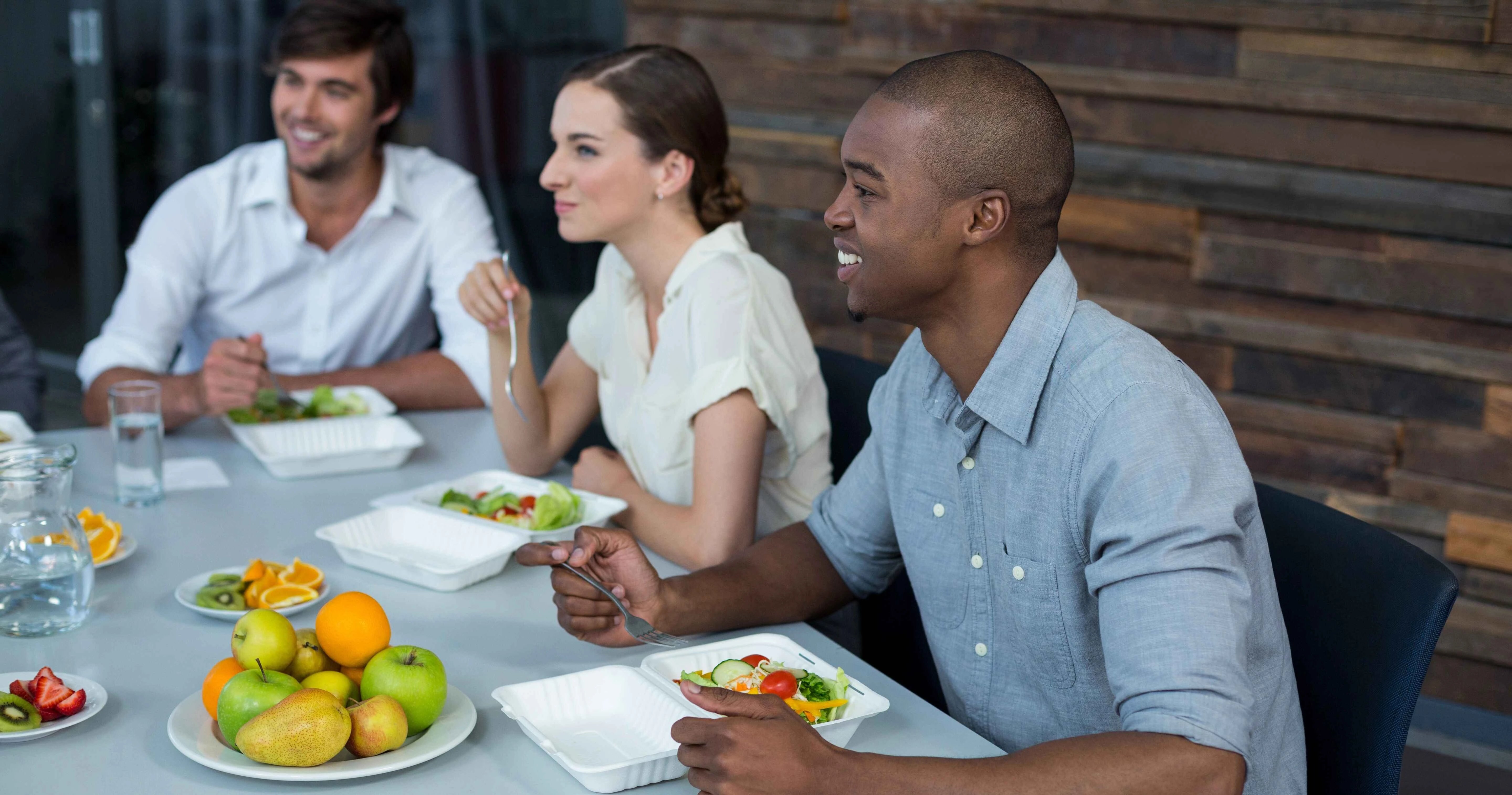 Three people sitting at a table eating healthy meals, with containers of salad and a plate of fruit including apples, oranges, and sliced kiwi.