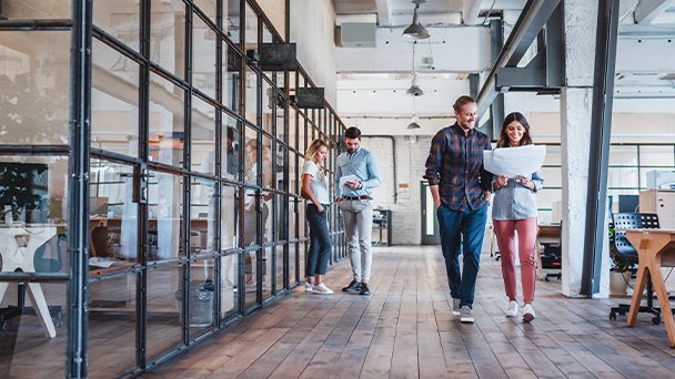 A diversed group of professionals walking through a modern office space, engaged in conversation and carrying laptops and notebooks