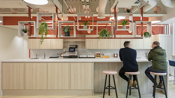 Two men sitting at a kitchen counter in an office, discussing work over coffee