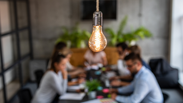 A light bulb from a string with people sitting and working at a desk in the background in blur