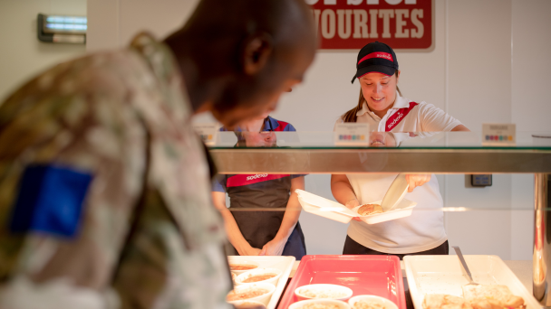 A woman in uniform serving food in the background with a male soldier in front of the frame