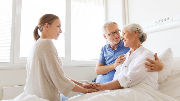 Female visitor chatting with senior female patient and her partner in hospital room
