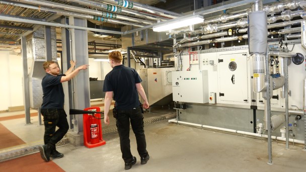 Two workers are standing in a room with pipes on the ceiling