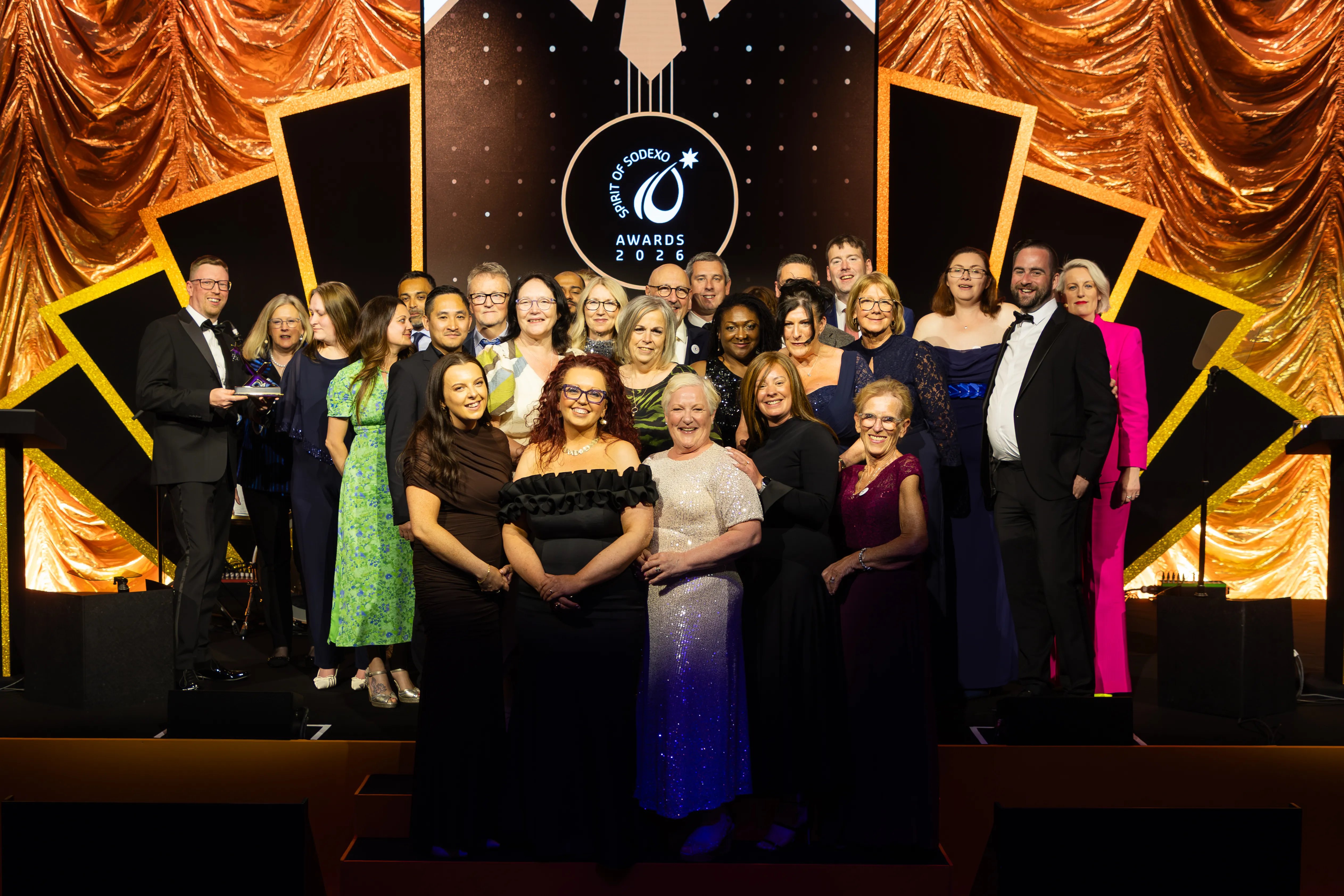 Group of award recipients and presenters posing on a stage in front of a gold curtain backdrop, with illuminated event branding displayed behind the group.