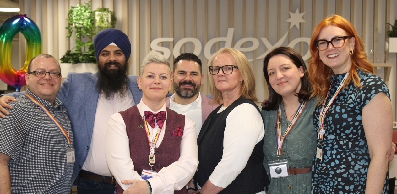 Group photo of seven Sodexo employees wearing rainbow lanyards during a workplace Pride celebration, standing together in front of a Sodexo branded backdrop with colourful anniversary decorations and plants in the office.