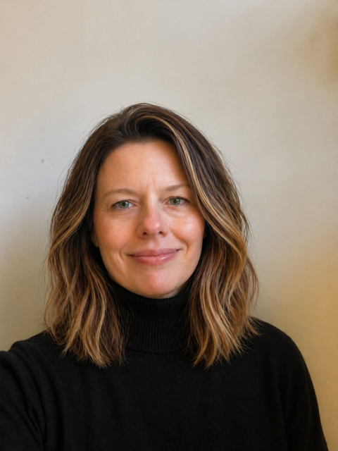 Portrait of a colleague standing against a plain light-coloured background, wearing a black long-sleeved top and dark trousers. The image is cropped from head to mid-thigh and features soft, natural lighting.