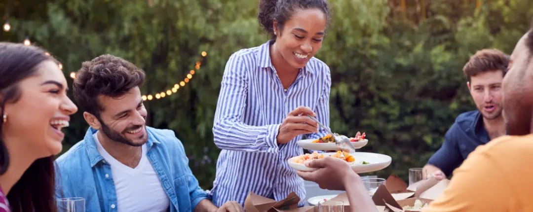 Group of people sharing food at an outdoor table, with one person serving a plate while others sit around in a garden setting with string lights.