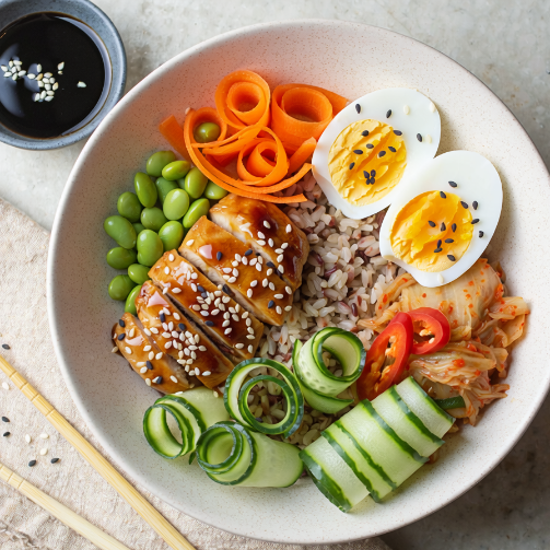  A bowl of sesame-glazed chicken with brown rice, edamame beans, sliced cucumber, carrot ribbons, kimchi, and two halved soft‑boiled eggs, with chopsticks beside the bowl.