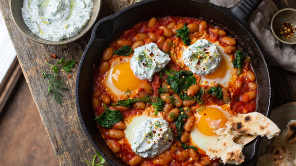 A cast‑iron pan filled with beans in spiced tomato sauce, topped with baked eggs, greens, dollops of yoghurt, and herbs, served with torn flatbread on a wooden surface.