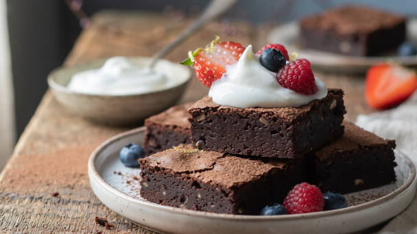 Three stacked slices of dark chocolate brownies on a ceramic plate, topped with a dollop of whipped cream and fresh berries, on a rustic wooden table.