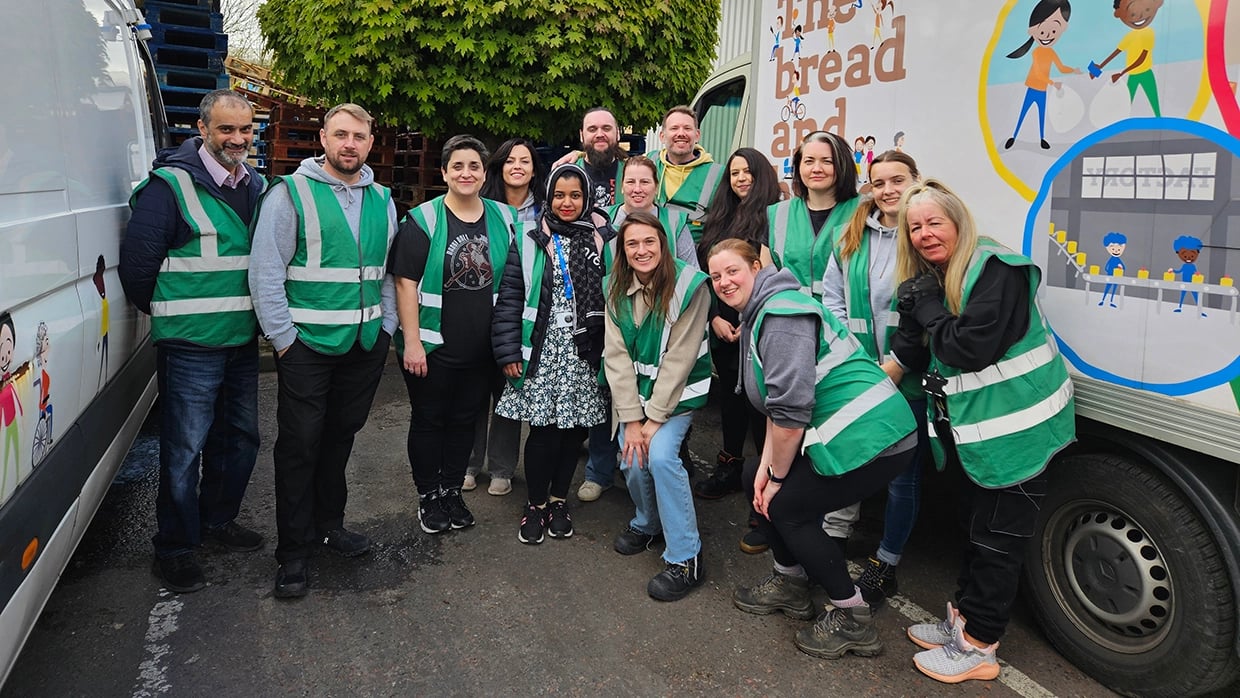 Group of volunteers wearing green high-visibility vests standing together outdoors next to a branded van, with stacked pallets and a leafy tree visible in the background.