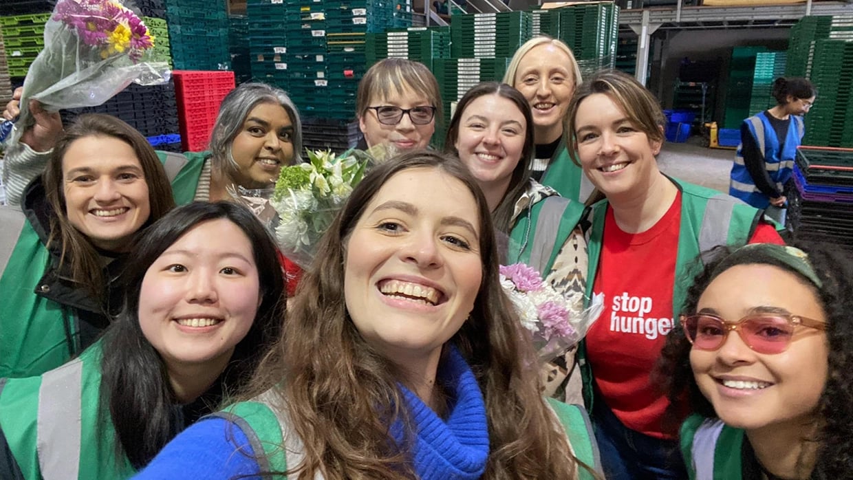 Group selfie of volunteers wearing green high-visibility vests inside a warehouse filled with stacked plastic crates, some holding flowers and one wearing a “Stop Hunger” t-shirt.