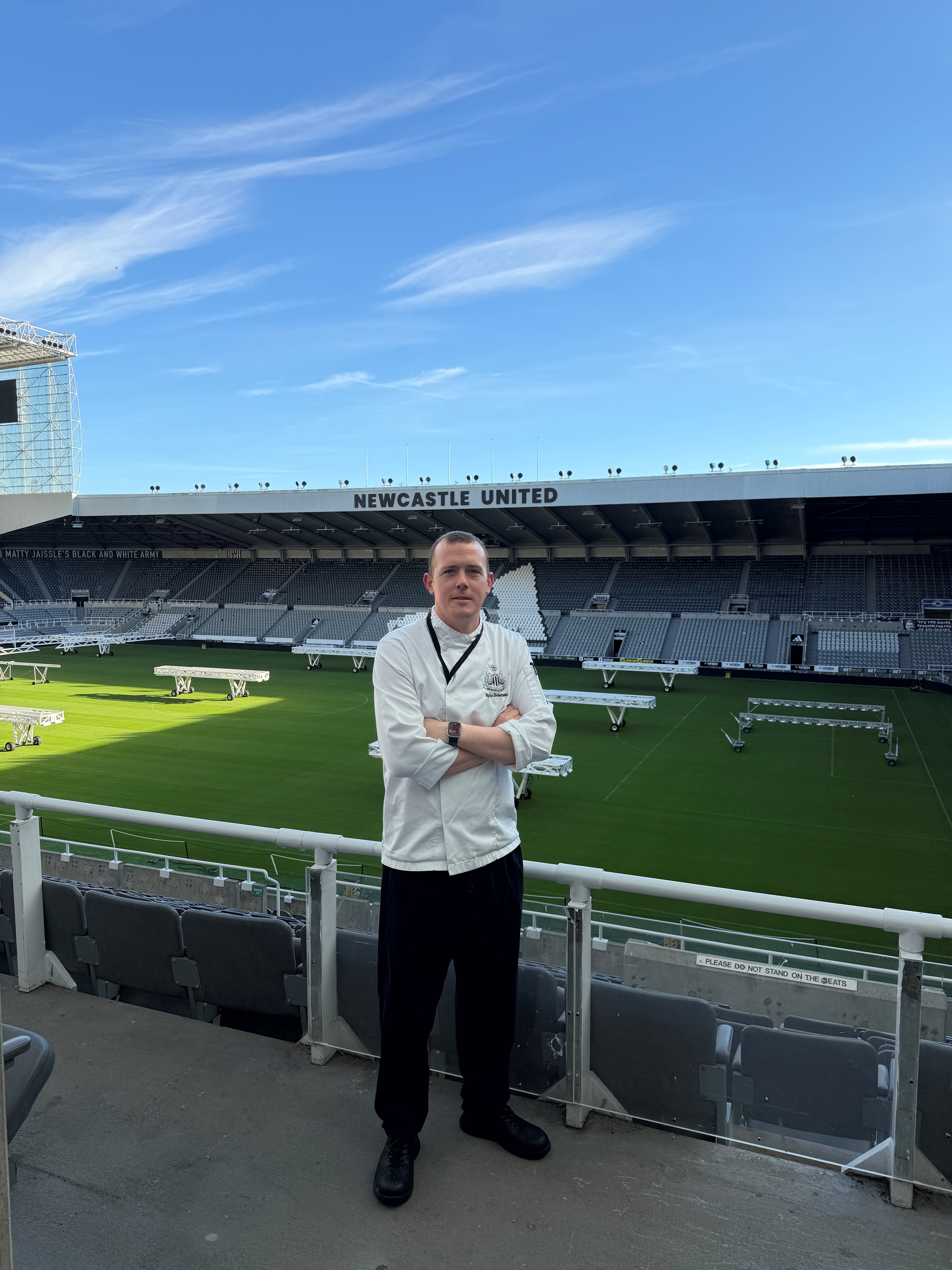 Person in catering uniform standing at a sports stadium overlooking the pitch.