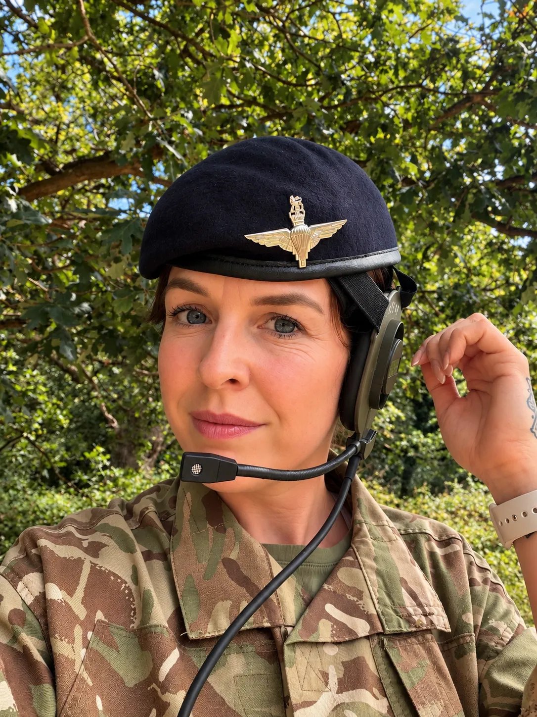Close-up portrait of a military service member wearing a camouflage uniform and dark beret with insignia. The individual is adjusting a headset earpiece attached to communication equipment. Trees and greenery are visible in the background of the outdoor setting.