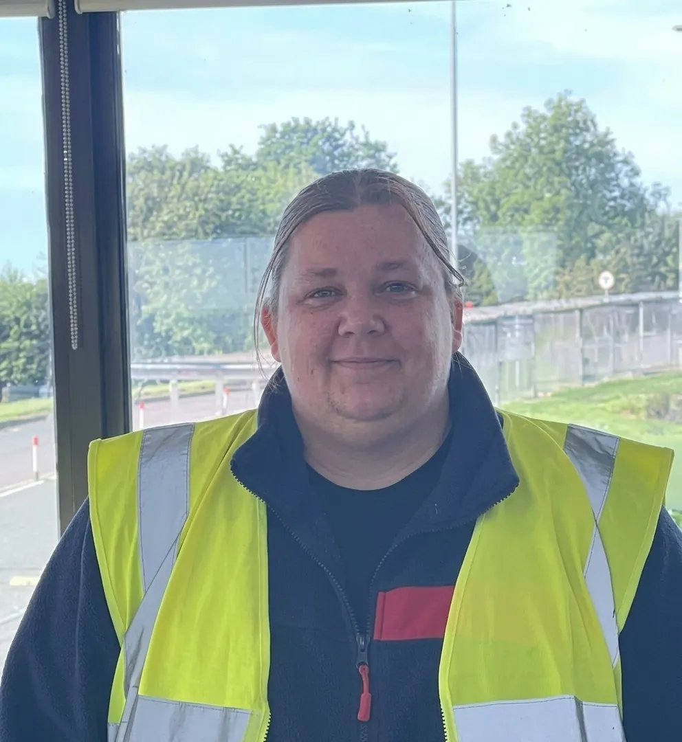 Sodexo colleague wearing a high-visibility vest standing indoors near large windows.