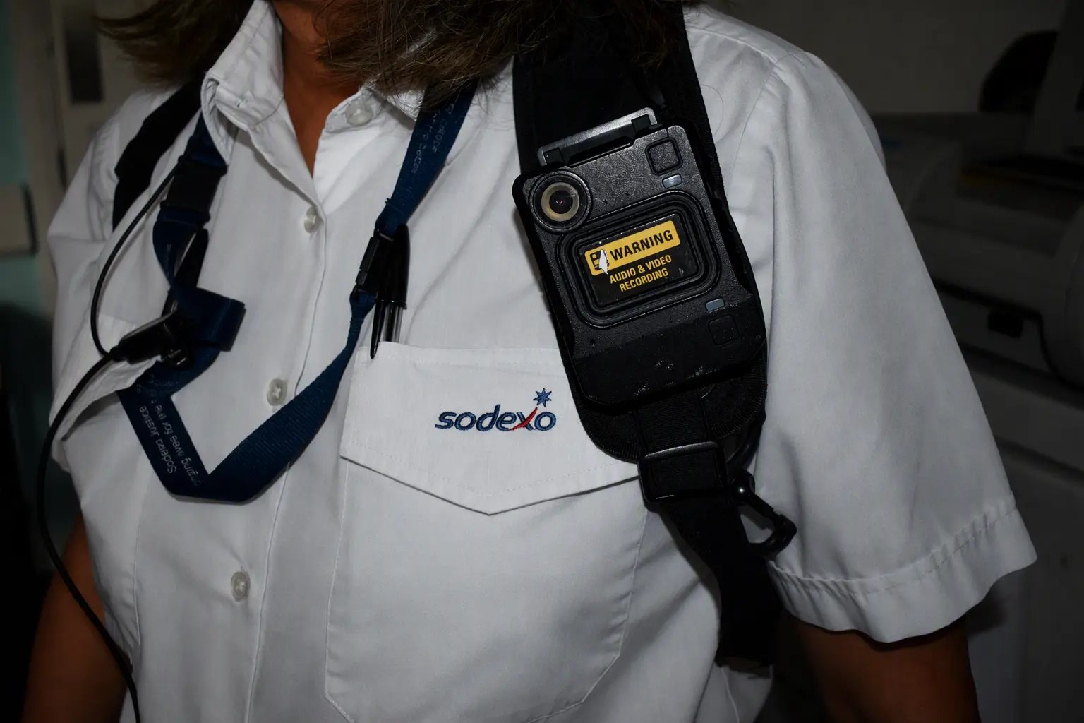 Close-up of a Sodexo employee uniform showing a body-worn camera attached to a shoulder strap. The white shirt features an embroidered Sodexo logo on the chest pocket, with a pen clipped inside the pocket. The camera has a visible warning label indicating audio and video recording.